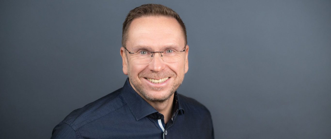 Mann mit Brille und kurzer Frisur, lächelt vor einem grauen Hintergrund.